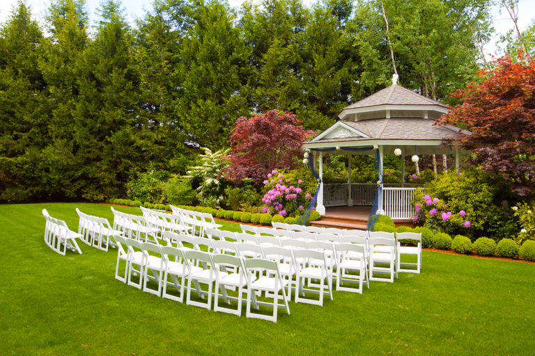 White folding chairs arranged on a lawn facing a garden gazebo before a ceremony