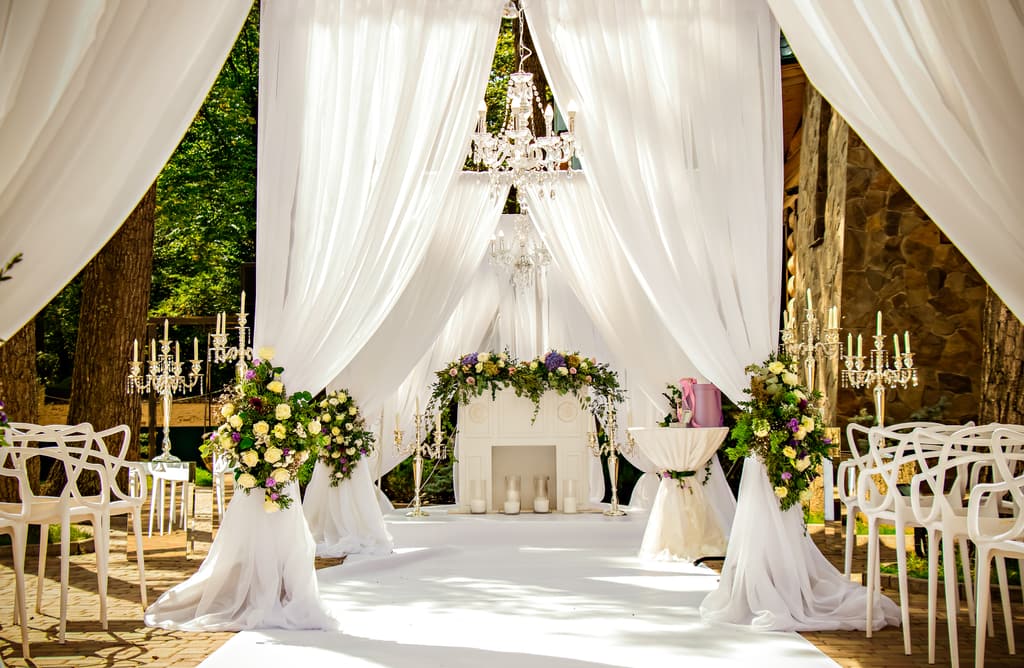 Outdoor wedding ceremony framed by white draped fabric, a chandelier, candelabras, and fresh floral arrangements